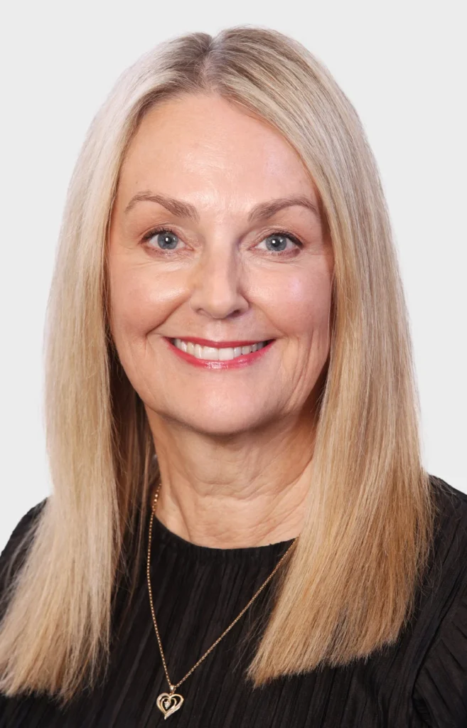Carole-reception - Anue Smiling head-and-shoulders portrait of a blonde woman wearing a black top and a gold heart necklace.