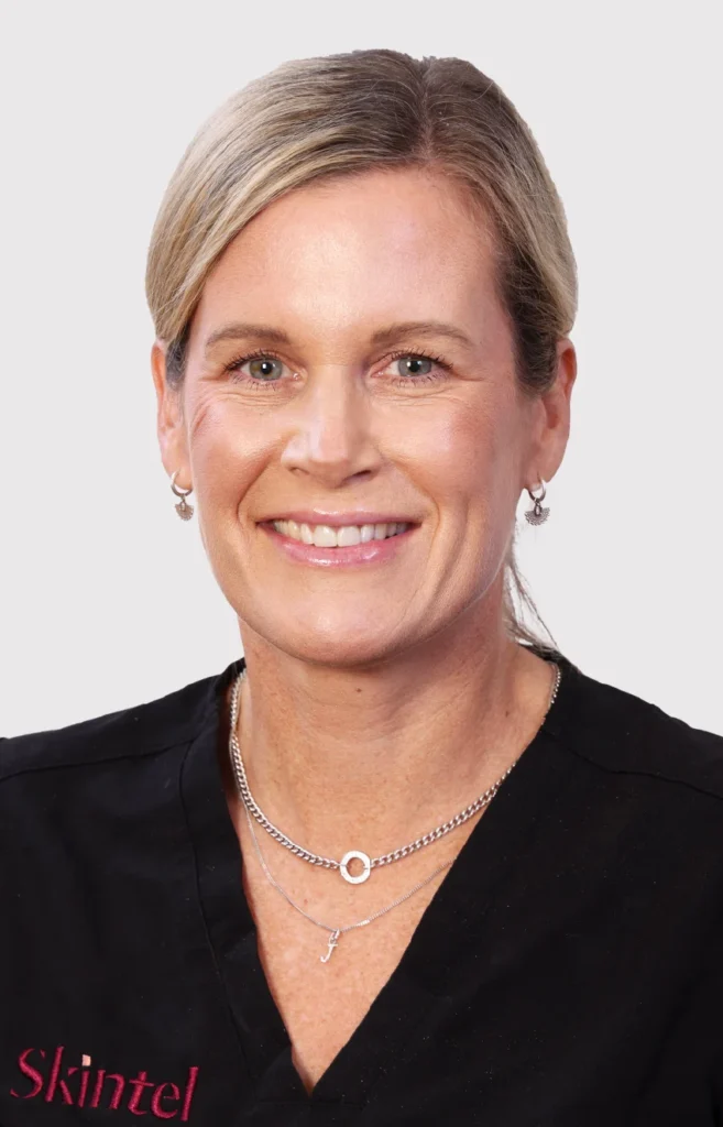 Jacinda-nurse - Anue Smiling blonde woman in black scrubs, wearing silver necklaces, against a light gray background.