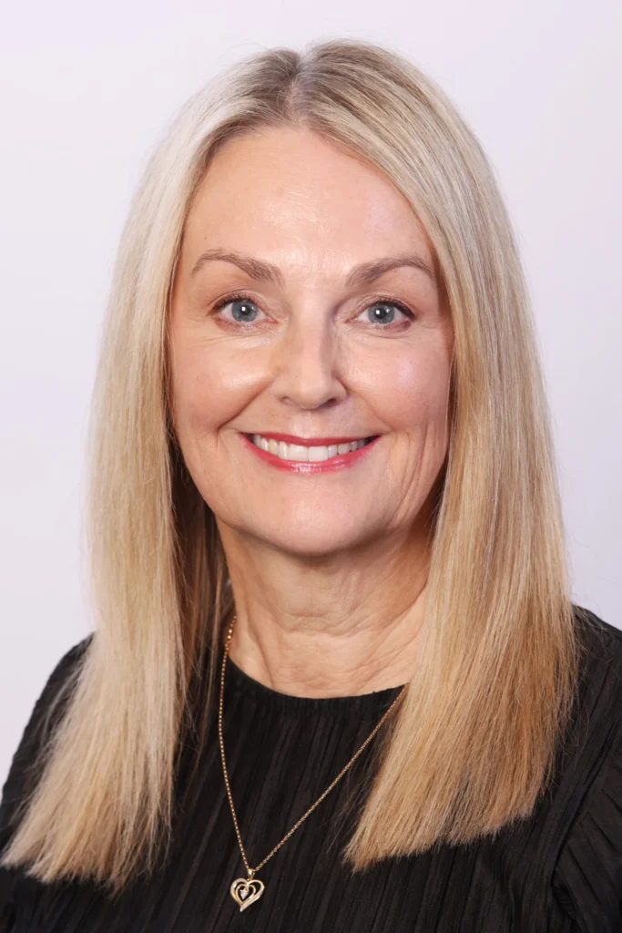 Smiling middle-aged woman with long blonde hair wearing a black top and a gold heart pendant necklace.