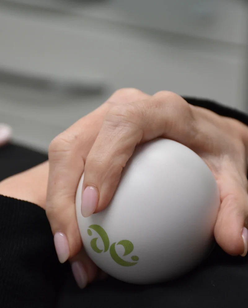 Hands clasped around a white egg-shaped stress ball with a green logo on a dark surface.